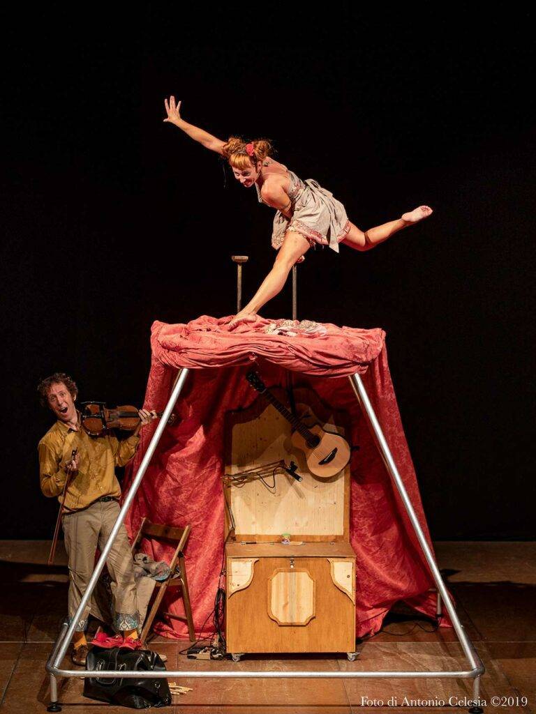 Foto spettacolo La dama Demodé, molti colori, donna in equilibrio sopra una struttura rossa tipo tenda, uomo con violino